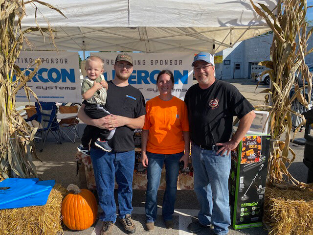 Four people, including a child, posing at the Nercon sponsor booth framed by cornstalks, hay bales and a pumpkin